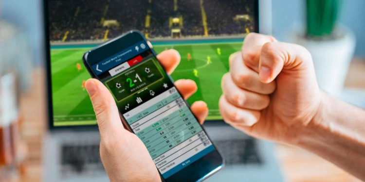 Persona con un celular en una apuesta celebrando un gol. Laptop al fondo, con un juego de fútbol.